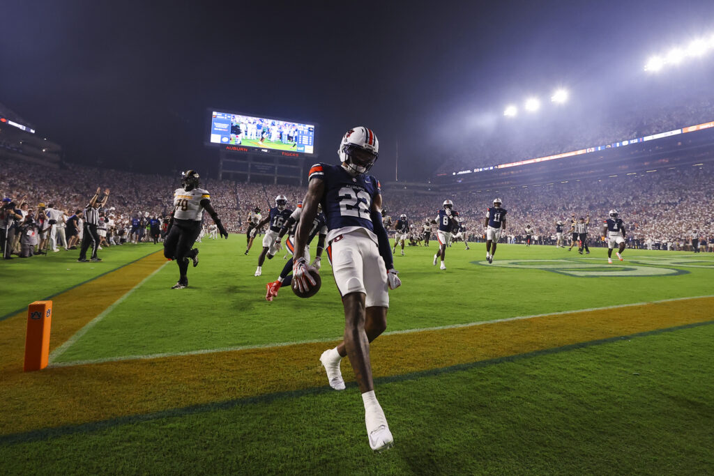 An athlete in a blue and white uniform runs across a football field at night, holding a ball, with other players and a large crowd in the background.