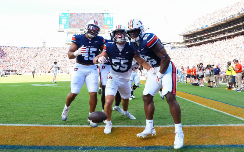 Three football players in blue and white uniforms celebrate on the field with a crowd in the stands behind them.